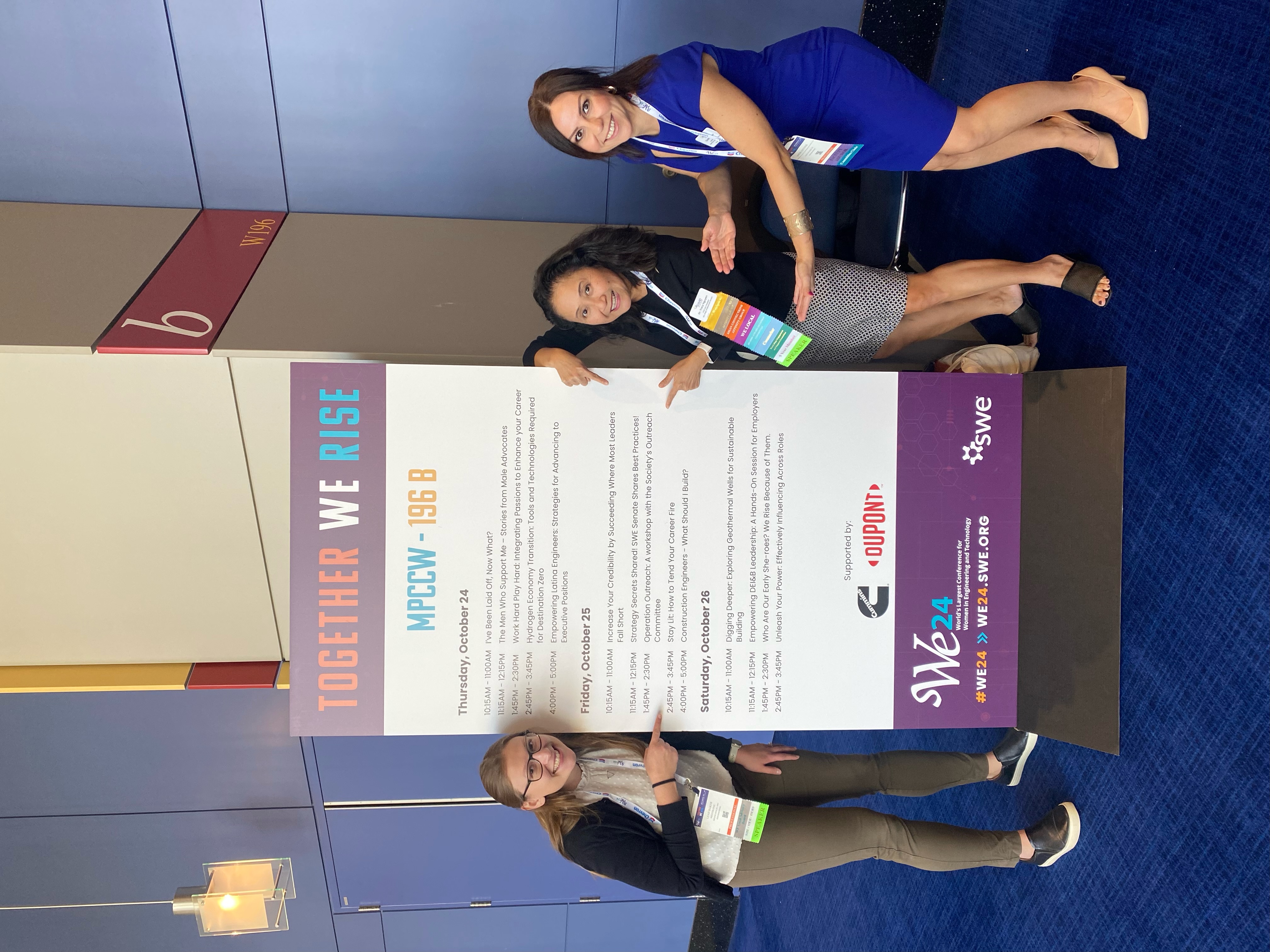 Leah stands with two fellow Society of Women Engineers members beside a WE24 conference schedule featuring sessions on outreach, leadership, and engineering.