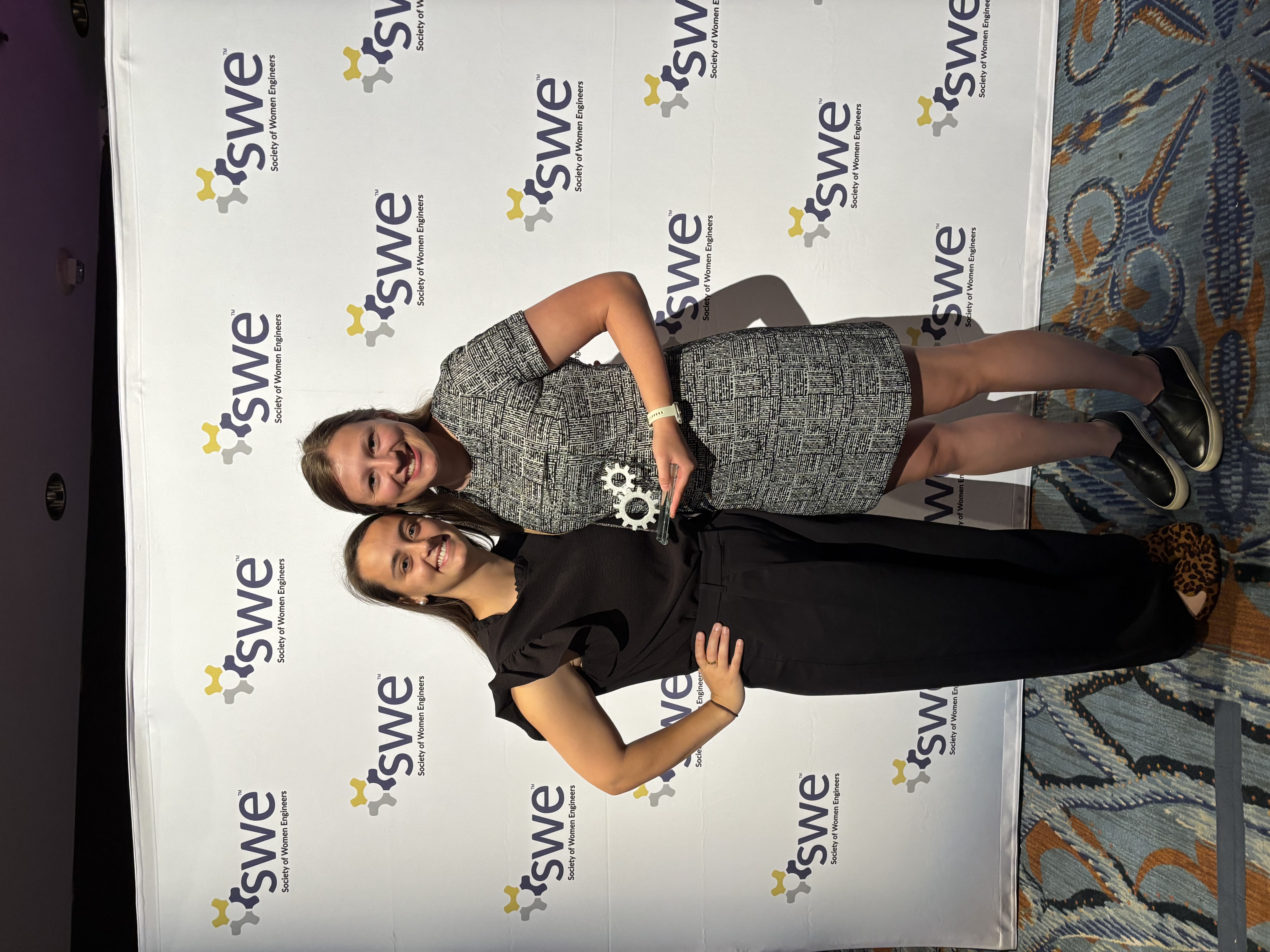 Leah stands with a colleague at a Society of Women Engineers event, smiling and holding an award in front of an SWE backdrop.