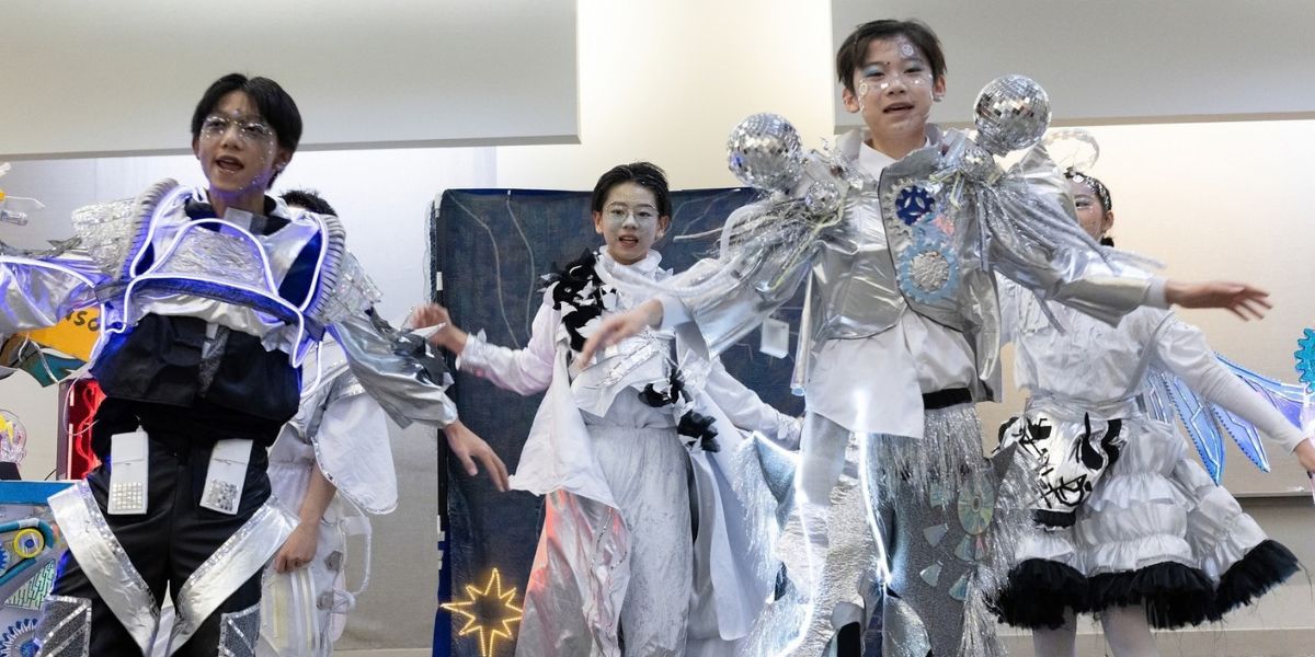 Destination Imagination students from China perform at Global Finals in futuristic silver, black, and white costumes with illuminated details, surrounded by colorful, student-created props and scenery.