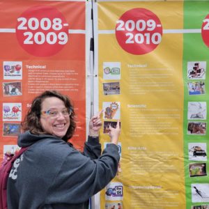 Andrew Keller stands in front of a large banner at Global Finals, displaying the year-to-year timeline of Destination Imaagination's Team Challenges. 