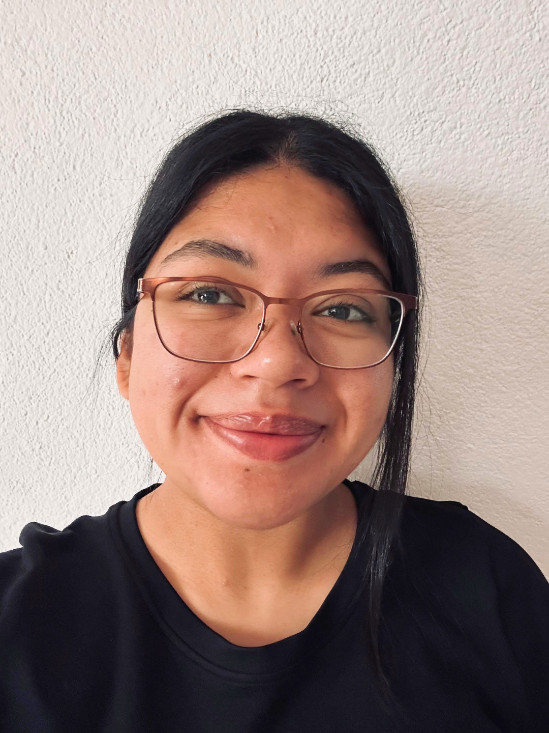 Dulce Ana Victoria Valencia Perez smiling in front of a light-colored wall. She is wearing glasses and a black shirt in a close-up portrait photo.