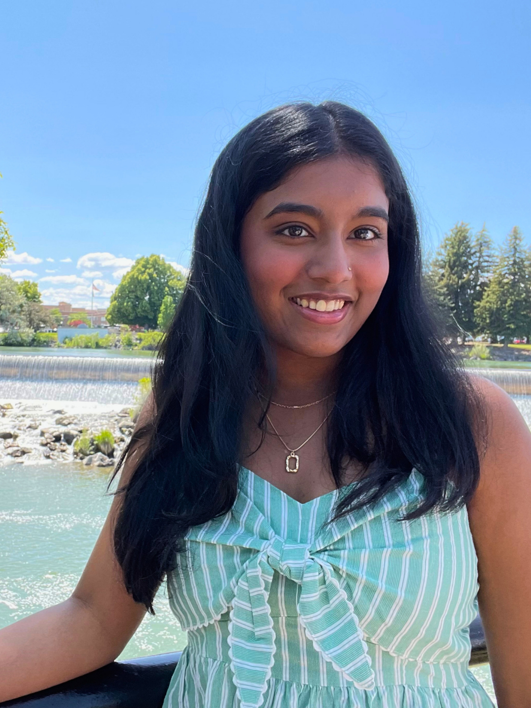 Pooja Rajesh smiling outdoors near a waterfront on a sunny day. She is wearing a light green and white striped dress and standing in front of trees, water, and a small waterfall.